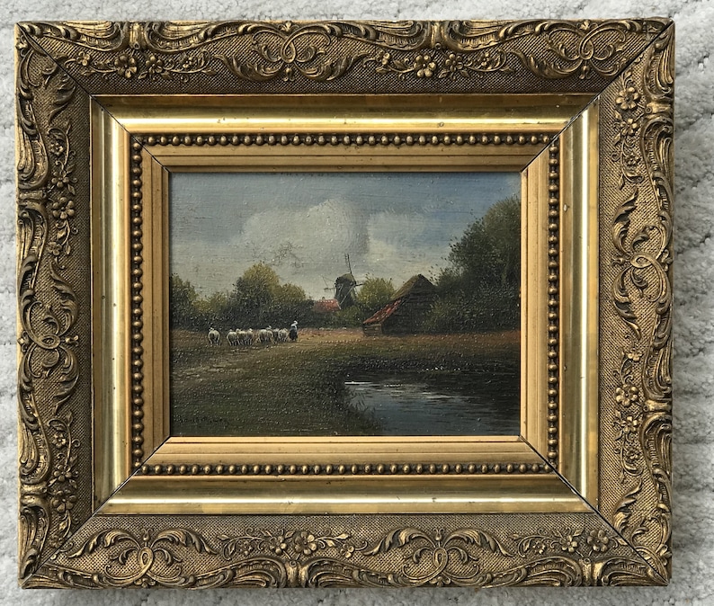 Op de afbeelding: Een ingelijst olieverfschilderij dat een landelijk landschap afbeeldt met een windmolen, een boerderij en een kudde schapen die grazen in een veld. Het schilderij is geplaatst tegen een bewolkte lucht en toont een klein beekje op de voorgrond.