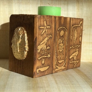 Könnte beinhalten: Kerzenhalter aus Holz mit in die Seiten geschnitzten ägyptischen Hieroglyphen. Der Halter ist braun und goldfarben bemalt und hat eine grüne Kerze im Inneren.