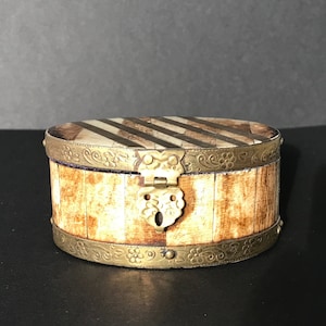 Könnte beinhalten: Eine kleine, runde, dekorative Schachtel aus Knochen mit einem Messingdeckel mit Scharnier und floralem Design. Die Schachtel hat ein gestreiftes Muster auf der Oberseite und eine Messingband um die Unterseite.