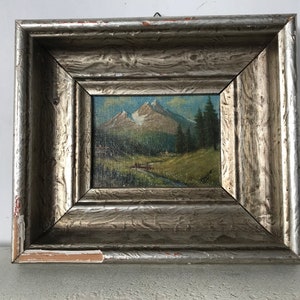 Könnte beinhalten: Ein gerahmtes Ölgemälde einer Berglandschaft mit einem kleinen Bach, der durch den Vordergrund fließt. Das Gemälde befindet sich in einem silbernen Rahmen mit einer abgenutzten Oberfläche.