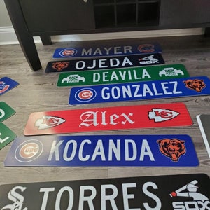 May include: A set of six personalized baseball nameplates with team logos. The nameplates are in various colors, including blue, red, green, and black. The logos include the Chicago Cubs, Chicago White Sox, Kansas City Chiefs, and Chicago Bears.