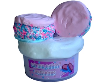Puede incluir: Un recipiente blanco de slime con una etiqueta rosa y azul que dice "Cotton Candy Macarons". Hay tres macarons rosas y azules encima del slime.