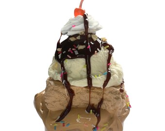 Puede incluir: Un sundae decorativo con capas de helado marrón y vainilla, cubierto con crema batida, salsa de chocolate, chispas y una cereza. La base del sundae tiene el texto "Fudge Brownie Sundae".