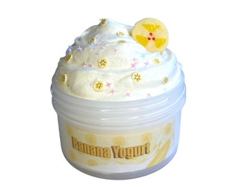 Può includere: Uno slime cremoso bianco con una fetta di banana gialla in cima, in un contenitore di plastica trasparente con un'etichetta che recita "Banana Yogurt".