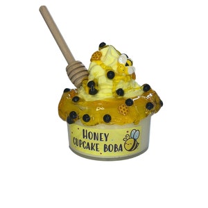 Op de afbeelding: Een gele honing cupcake boba met zwarte boba parels en honing besprenkeld, bedekt met slagroom en een honinglepeltje. De verpakking heeft een label met de tekst "Honey Cupcake Boba" met een cartoon-bij.