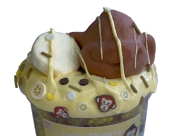 Puede incluir: Un slime amarillo con una cabeza de mono marrón en la parte superior, decorado con un chorrito de chocolate blanco, espolvoreado blanco y amarillo y pequeños amuletos de mono. El slime está en un recipiente transparente con una etiqueta que dice "Funky Monkey Crunch".