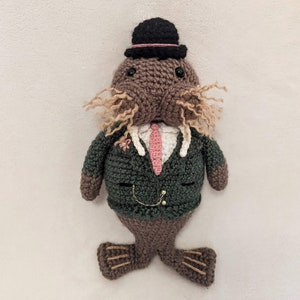 May include: A crocheted walrus wearing a green suit, a pink tie, and a black top hat. The walrus has brown fur and a long, brown mustache.