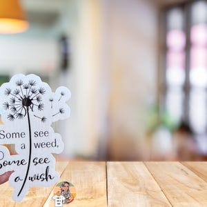 May include: A white sticker with a black dandelion design and the text "Some see a weed, Some see a wish".