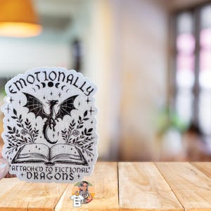 May include: A white sticker with black text and a black and white illustration of a dragon. The text reads "Emotionally Attached to Fictional Dragons".