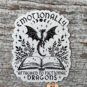 May include: Black and white sticker with a dragon, book, and the text "Emotionally Attached to Fictional Dragons".