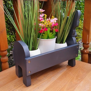 May include: Black wooden planter with three white pots filled with green plants and pink flowers. The planter is shaped like a dog and has a dark wood finish.
