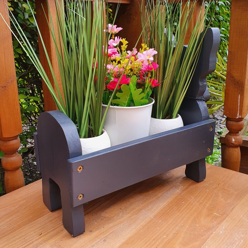 May include: Black wooden planter with three white pots filled with green plants and pink flowers. The planter is shaped like a dog and has a dark wood finish.