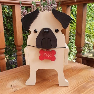 May include: A white wooden planter shaped like a pug dog with black ears and a black nose. The planter has a red bone-shaped tag with the name "Fred" written on it.