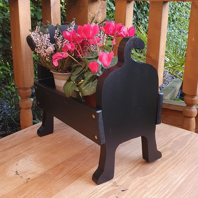 May include: Black wooden planter shaped like a dog with pink and white flowers in pots. The planter has a unique design with a curved back and legs.