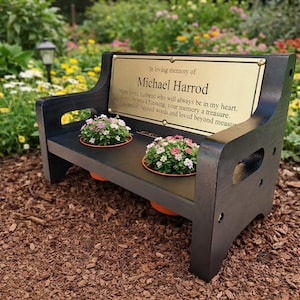 May include: A black wooden memorial bench featuring a gold-toned metal plaque with the text In loving memory of Michael Harrod, Much loved husband who will always be in my heart, Your life was a blessing, your memory a treasure, You are missed beyond words and loved beyond measure. Two orange pots with pink and white flowers are set into the bench seat.