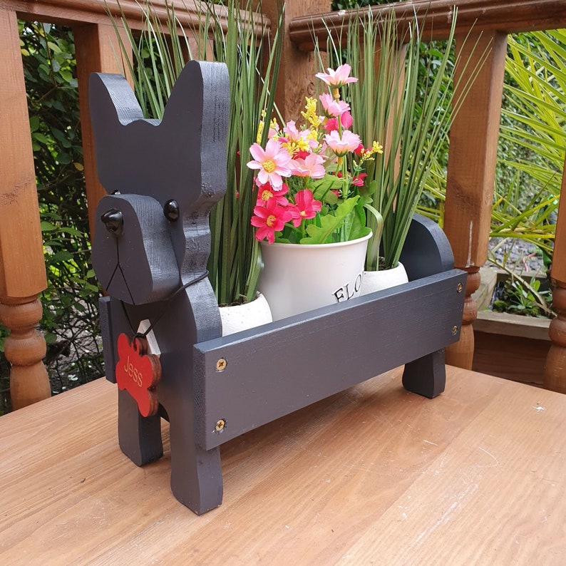 May include: Black wooden dog planter with a red paw print tag that says "Jess" and two white pots with green plants and pink flowers. The planter is on a wooden surface.