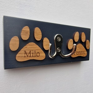 May include: A navy blue wooden pet leash holder with two paw print cutouts and the names "Milo" and "Penny" engraved on them. A silver metal hook is attached to the board.