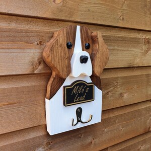 May include: Wooden dog head shaped coat rack with a white and brown painted finish. The dog's nose is black and the eyes are black with white pupils. The rack has a black sign that reads "Milo's Lead" and two silver hooks.