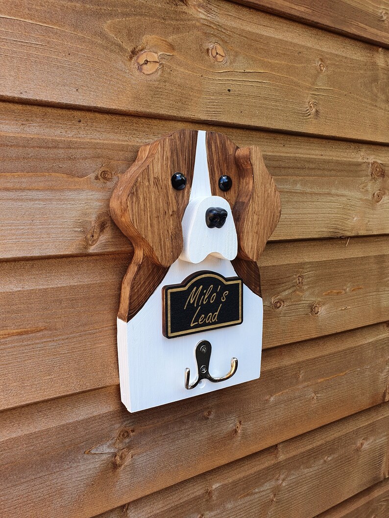 May include: Wooden dog head shaped coat rack with a white and brown painted finish. The dog's nose is black and the eyes are black with white pupils. The rack has a black sign that reads "Milo's Lead" and two silver hooks.