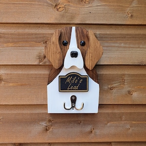 May include: Wooden dog-shaped key holder with a white and brown painted finish. The dog has black eyes and a black nose. The holder has a black sign that reads "Milo's Lead" and two metal hooks.