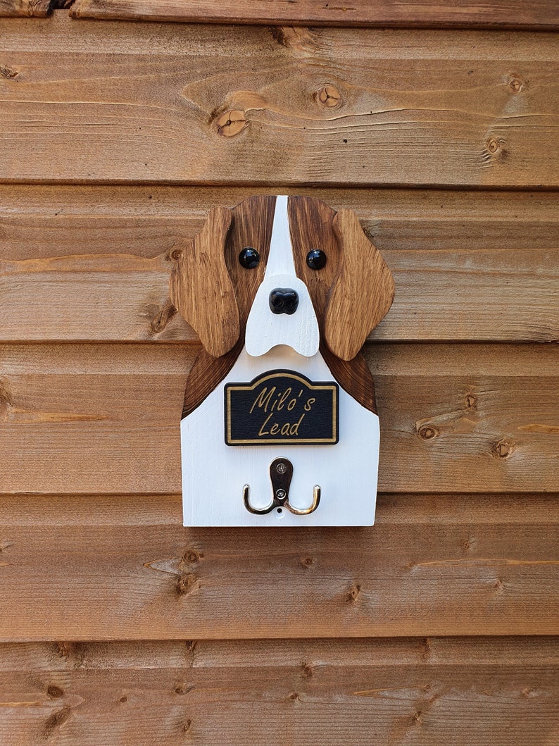 May include: Wooden dog-shaped key holder with a white and brown painted finish. The dog has black eyes and a black nose. The holder has a black sign that reads "Milo's Lead" and two metal hooks.