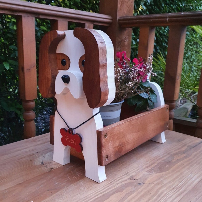 Op de afbeelding: Houten plantenbak in de vorm van een hond met bruine oren en een wit lichaam. De plantenbak heeft een rood botvormig label met de naam "Fred" erop. De plantenbak is gevuld met een kleine plant in een pot met roze bloemen.