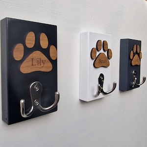 Può includere: Tre ganci in legno per guinzaglio per cani con disegni di impronte di zampe. I ganci sono dipinti di nero, bianco e grigio. Ogni gancio ha un gancio doppio e un'impronta di zampa con un nome inciso su di esso. I nomi sono Lily, Benny e Lily.