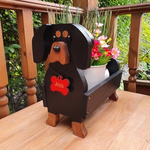 May include: Black wooden dachshund planter with a red bone tag that says "Jess" and a white pot with green plants and pink flowers.