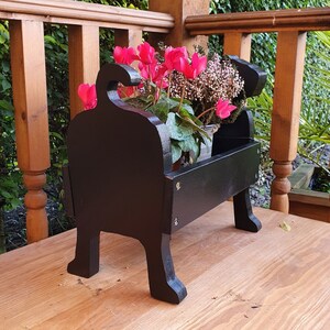 May include: Black wooden planter shaped like a dog, holding pink cyclamen flowers in a pot. The planter is sitting on a wooden surface.