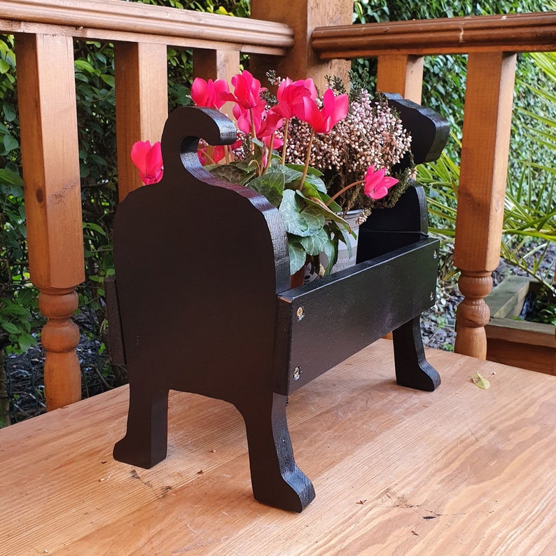 May include: Black wooden planter shaped like a dog, holding pink cyclamen flowers in a pot. The planter is sitting on a wooden surface.