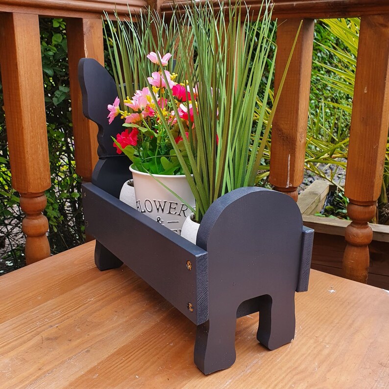 May include: Black wooden planter with two compartments, featuring a white ceramic pot with pink and white flowers and the word "FLOWER" printed on the side.