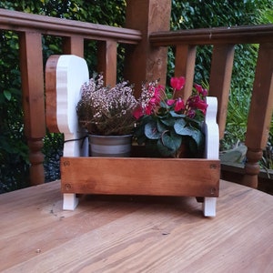 Op de afbeelding: Een houten plantenbak in de vorm van een hond, wit geschilderd, met twee potplanten. De plantenbak heeft een bruine houten doos met een witte rand. De planten zijn een roze cyclamen en een witte heide.