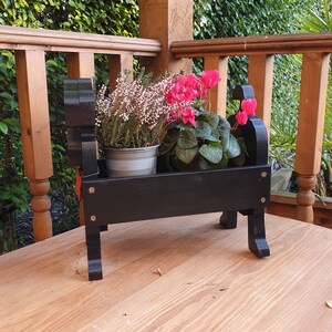 May include: Black wooden planter shaped like a dog, with two pink flowering plants inside. The planter is sitting on a wooden surface.