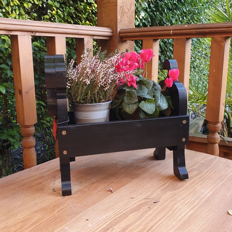 May include: Black wooden planter shaped like a dog, with two pink flowering plants inside. The planter is sitting on a wooden surface.