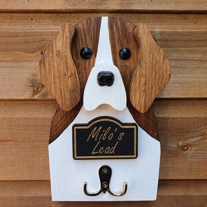 May include: Wooden dog-shaped wall hook with a white and brown paint finish. The hook has a black sign that reads "Milo's Lead" and two silver hooks.