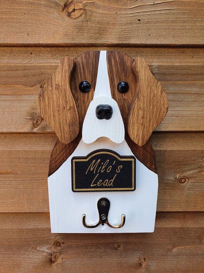 May include: Wooden dog-shaped wall hook with a white and brown paint finish. The hook has a black sign that reads "Milo's Lead" and two silver hooks.