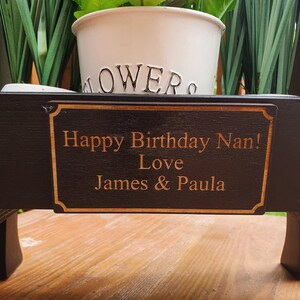 May include: A black wooden planter with a personalized plaque that reads "Happy Birthday Nan! Love James & Paula". The planter has a white pot with green plants inside.