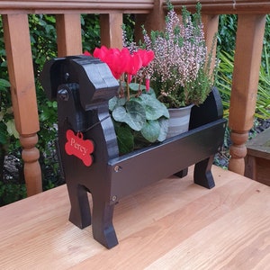 Könnte beinhalten: Schwarzer Holz-Hunde-Pflanztopf mit einem roten Schild, auf dem "Percy" steht, und einem Topf mit rosa und weißen Blumen.