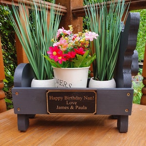 May include: A black wooden planter with three pots, two with green plants and one with pink and red flowers. The planter has a personalized plaque that reads "Happy Birthday Nan! Love James & Paula".