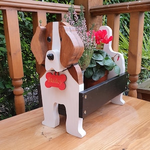 May include: Wooden dog planter with a black planter box. The dog is painted white with brown ears and a brown nose. The dog has a red bone tag with the name "Bailey" on it.