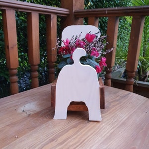 Op de afbeelding: Witte houten plantenbak in de vorm van een persoon met een roze en witte bloemschikking erin. De plantenbak staat op een houten oppervlak.