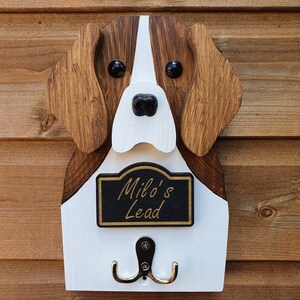 May include: Wooden dog head shaped wall hook with a white and brown painted finish. The dog has black eyes and a black nose. The hook has a black sign that reads "Milo's Lead".