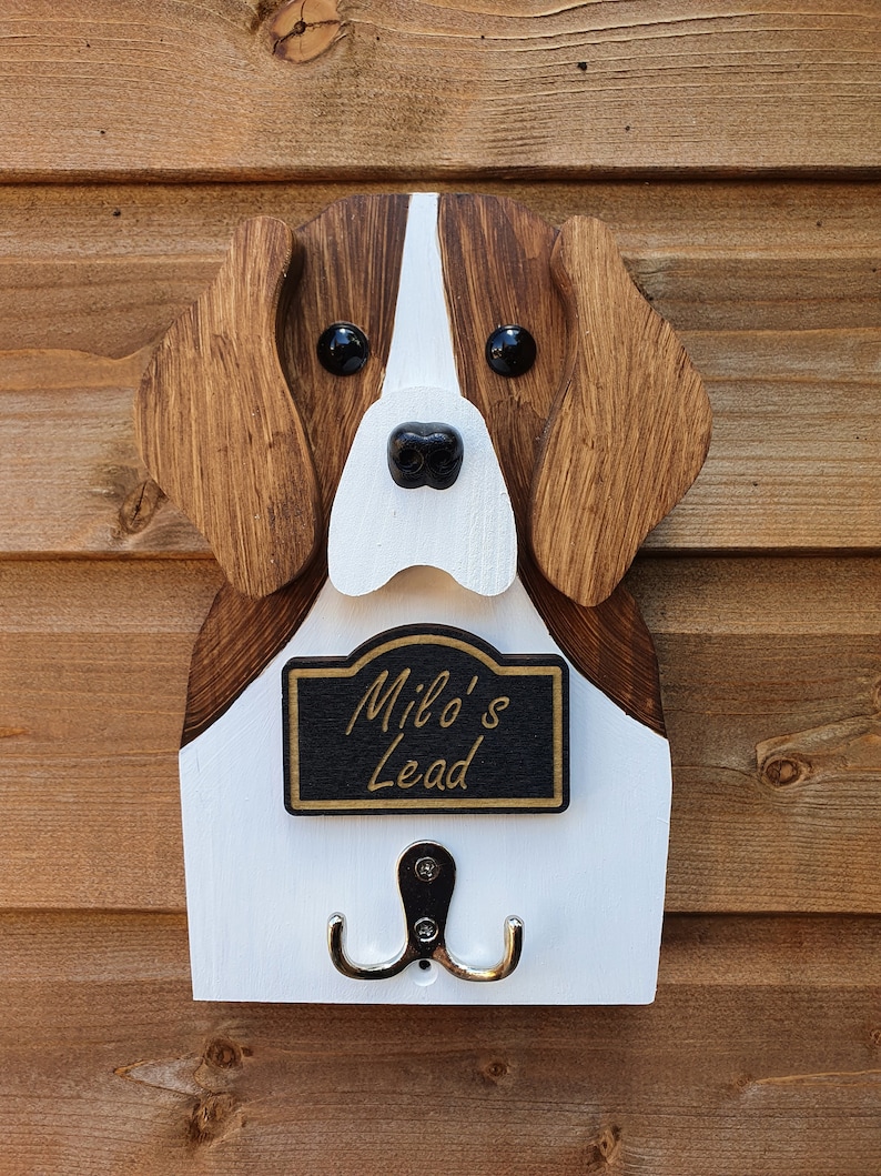 May include: Wooden dog head shaped wall hook with a white and brown painted finish. The dog has black eyes and a black nose. The hook has a black sign that reads "Milo's Lead".