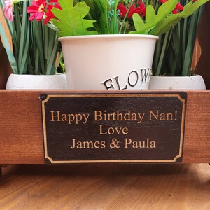 Op de afbeelding: Een houten plantenbak met een gepersonaliseerd bord met de tekst "Happy Birthday Nan! Love James & Paula". De plantenbak is gevuld met groene planten en roze bloemen.