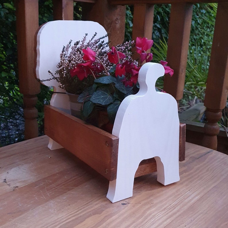 Op de afbeelding: Witte houten plantenbak in de vorm van een bed met een bruine houten basis. De plantenbak is gevuld met roze en rode bloemen.