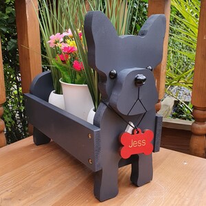 May include: Black wooden planter shaped like a French bulldog with a red bone-shaped tag that says "Jess". The planter is filled with green plants and flowers.