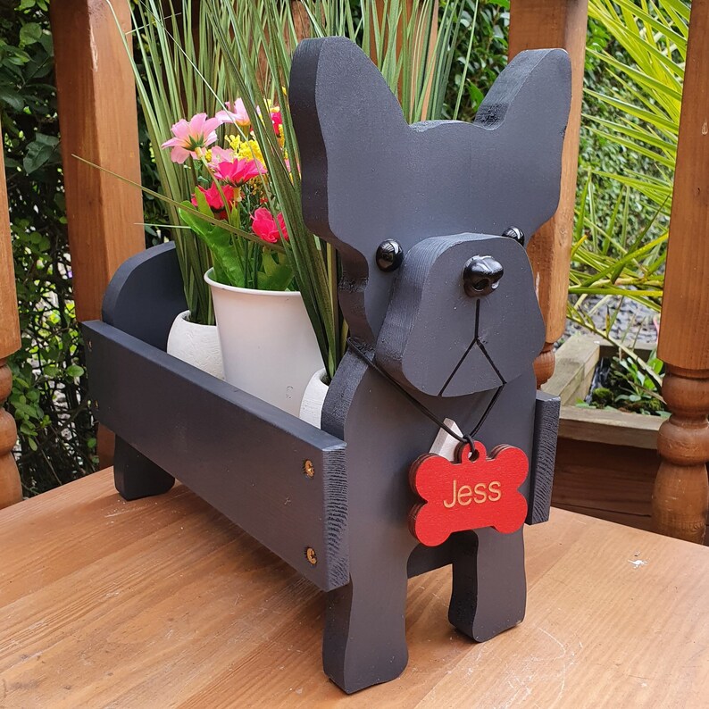 May include: Black wooden planter shaped like a French bulldog with a red bone-shaped tag that says "Jess". The planter is filled with green plants and flowers.