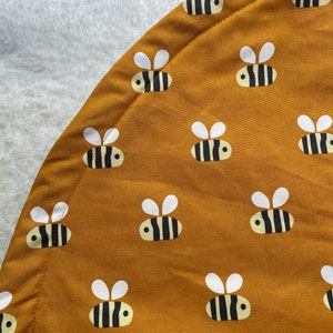 May include: Mustard-coloured, circular play mat featuring a repeating pattern of cartoon bees. Each bee has a yellow body with black stripes and white wings. The mat is bordered with a stitched seam.