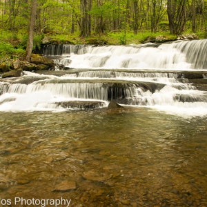 May include: A cascading waterfall flows over a series of rocky steps, creating a tranquil scene in a lush green forest.