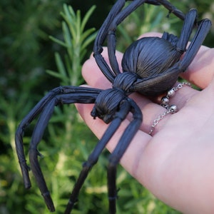May include: A black spider made of woven straw, held in a hand against a green leafy background.